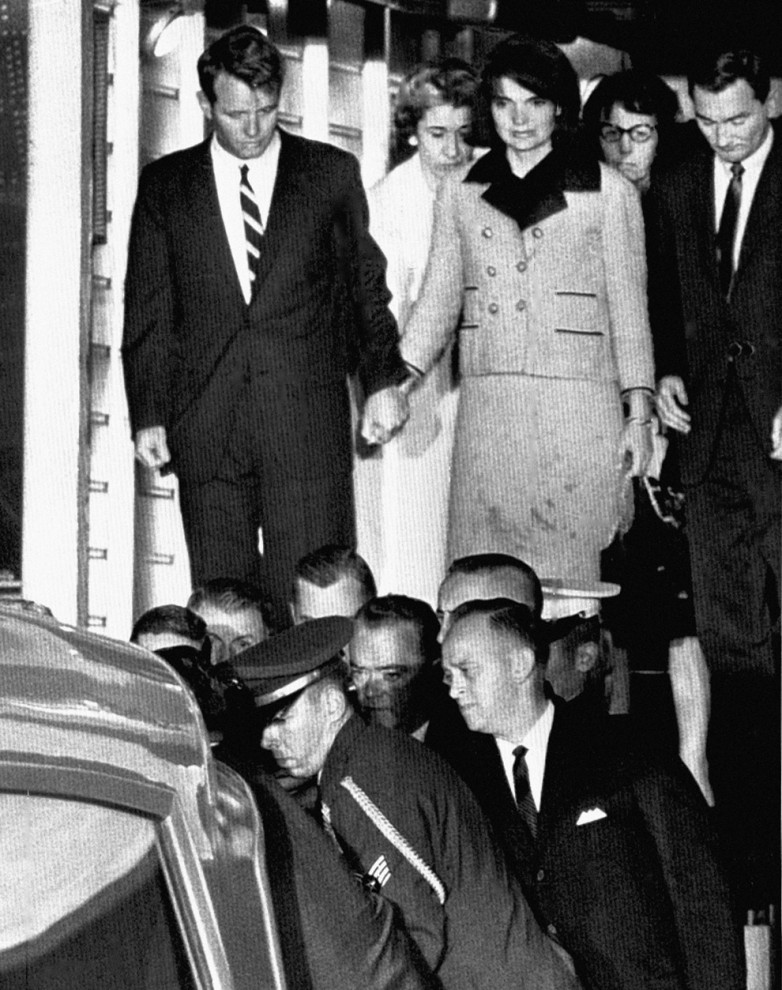 Роберт Кеннеди держит за руку Жаклин Кеннеди, пока гроб с телом Джона Кеннеди помещают в машину скорой помощи, по прибытии на авиабазу Эндрюс в штате Мэриленд. На одежде Жаклин Кеннеди видны пятна крови. (Associated Press)