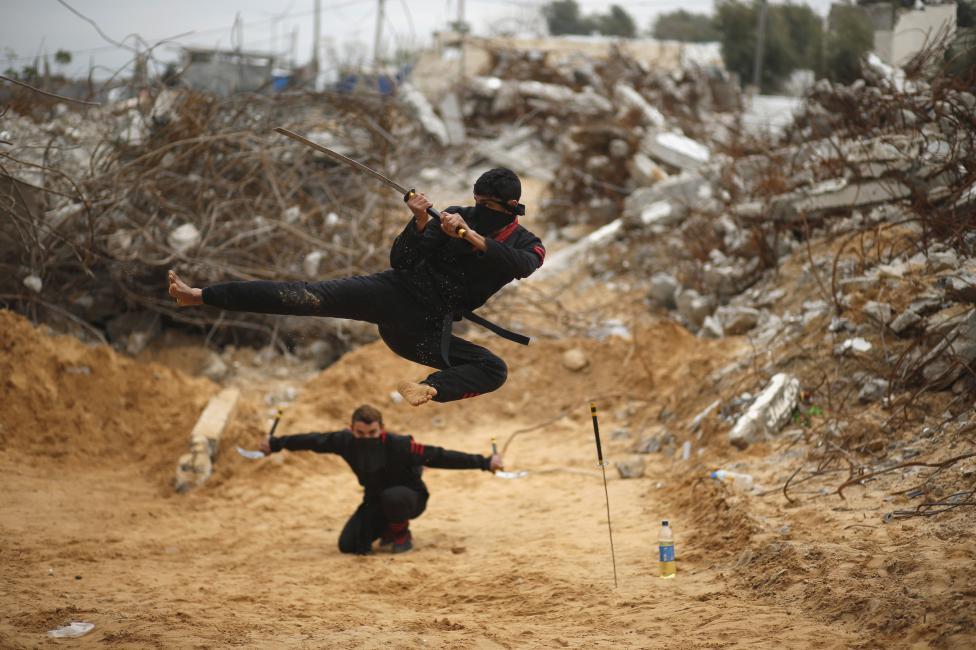 Палестинские подростки изображаюют ниндзя перед объективом фотографа на фоне зданий, разрушенных во время войны в 2014 году, в секторе Газа, 29 января 2016 года.